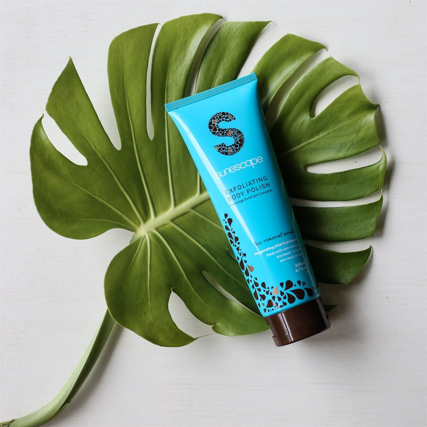 Blue tube of Sunescape Exfoliating Body Polish on a large green leaf with a white background