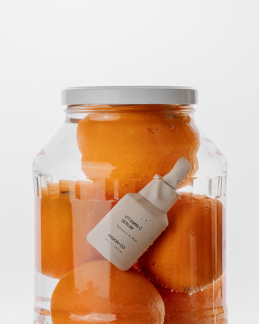 Jar of oranges with a vitamin C serum bottle inside on a white background