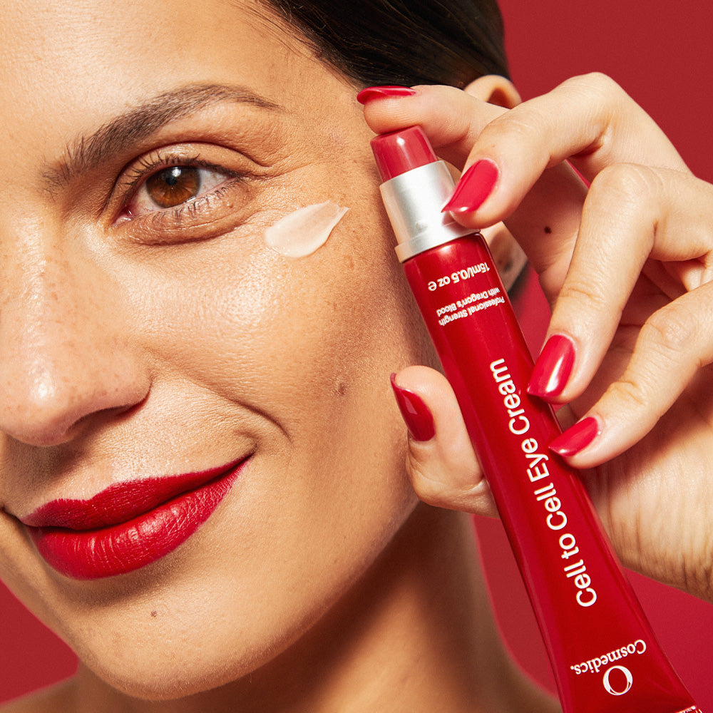 Woman applying 'O' Cell to Cell Eye Cream to her face with a red background