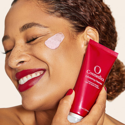 Woman applying 'O' Exfoliating Cleanser to face with pink patch, against a neutral background