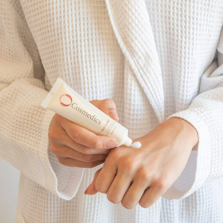 Person applying cream from a O Cosmedics Medi Soothe on their wrist, wearing a beige robe.