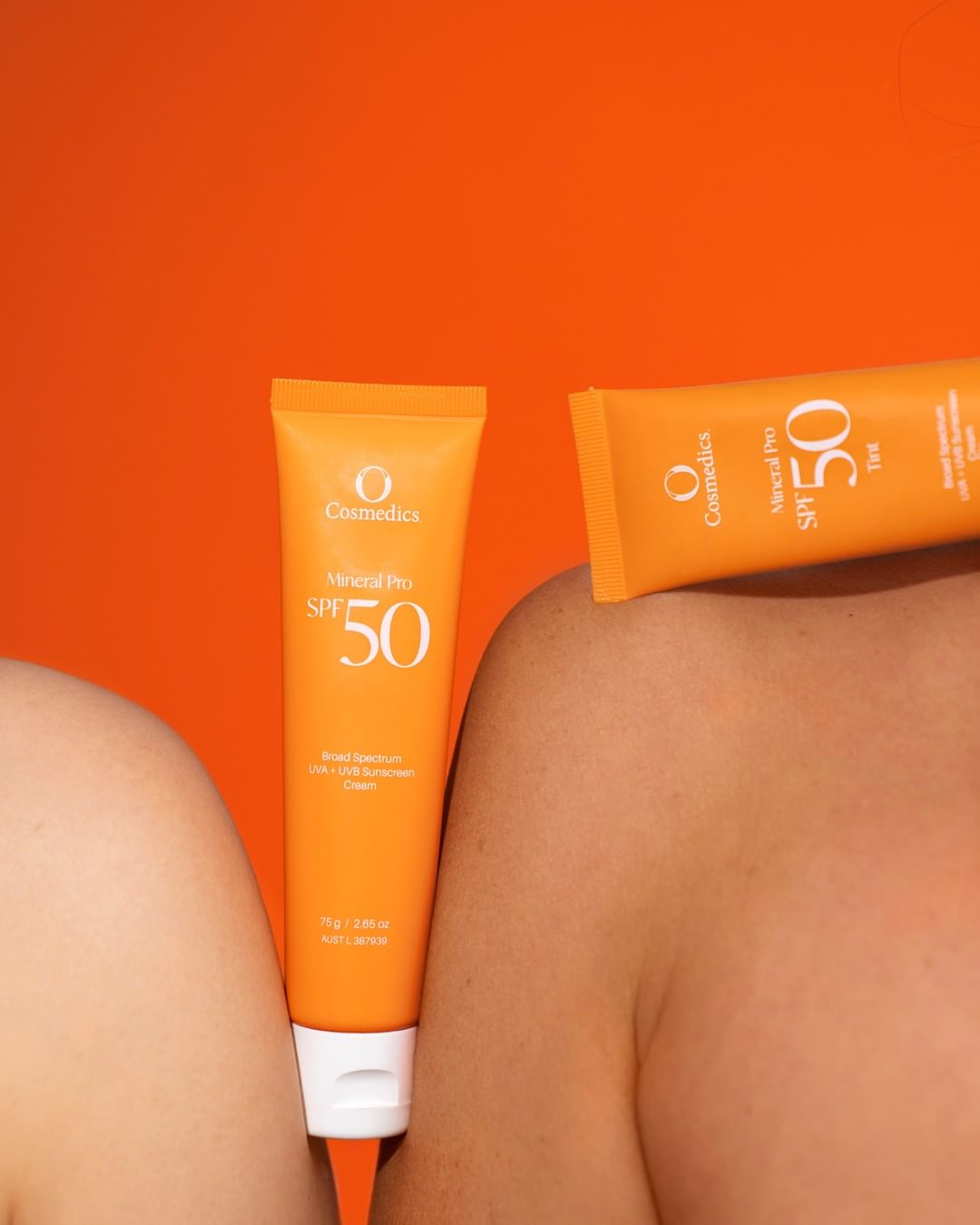 Two tubes of OCosmoetics Sunscreen SPF 50 on a person's shoulder against an orange background