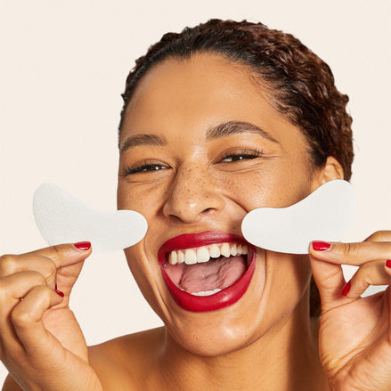 Woman holding 'O' No Baggage Native Collagen Eye Masks in front of her face against a beige background