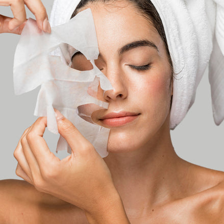 Woman applying O Cosmedics V8 Peptide Complex Mask  with a towel on her head against a neutral background