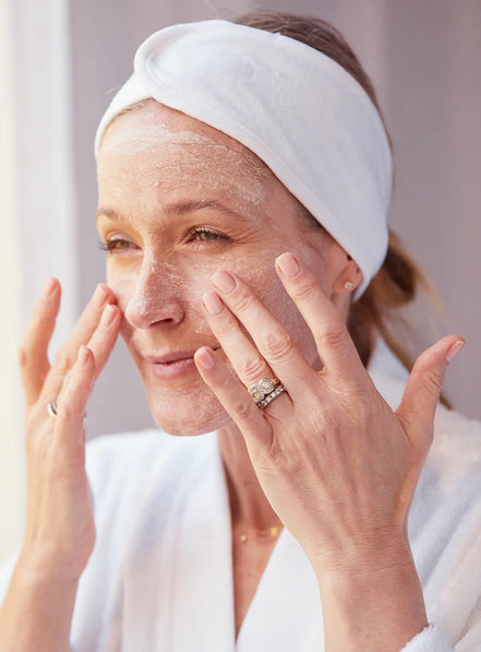Woman applying Anti Ageing 3 Fruit Enzymes Exfoliating Scrub & Mask to her face with a white towel on her head, wearing a white robe.
